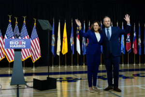 Kamala Harris and Douglas Emhoff wave on stage by a Biden-Harris podium.