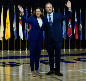 Kamala Harris and Douglas Emhoff wave on stage by a Biden-Harris podium.