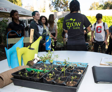 A group of people standing behind a table with plants and gardening supplies on it. One person has the words "Let's Grow Together" on the back of her shirt