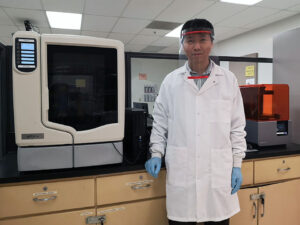 Assistant professor Bingbing Li stands next to a 3-D printer, wearing a face shield created in his lab.