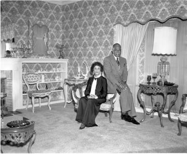An African American couple, seated, pose for a portrait in a well appointed room inside of a home.