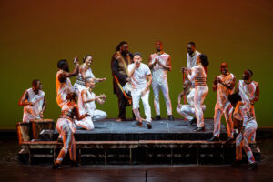 A singer surrounded by clapping dancers in African-inspired costumes and one drummer.