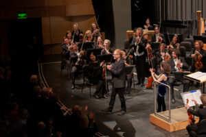 Composer Danny Elfman clasps his hands as the crowd applauds. He stands on The Soraya stage with violinist Sandy Cameron and the Royal Scottish National Orchestra.