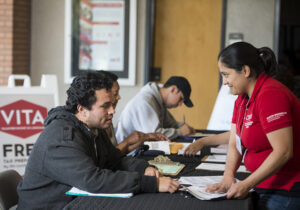 A man, seated, speaks with a woman who is across the table from him. She wears a red polo shirt with branding that identifying her as the VITA volunteer. They discuss a document that is on the table. In the background, another man is filling out paperwork.