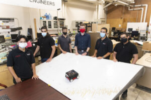 A group of students wearing masks standing up around a white table where there is a robot at the center.