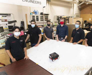A group of students wearing masks standing up around a white table where there is a robot at the center.