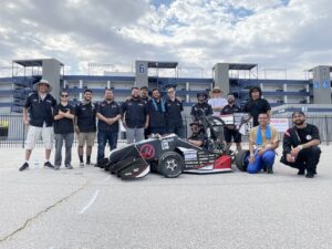 The students and the race car are smiling for the picture, standing in front of a building.