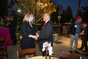 Exterior evening. President Beck, wearing a surgical face mask, speaks with Milt Valera. Both are standing and Milt clasps President Becks hands in a gesture of warmth.