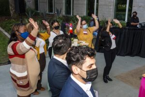 A group of 5 women and 2 men listen to the speaker (out of frame). The women raise both hands to the air, showing support for the speaker.