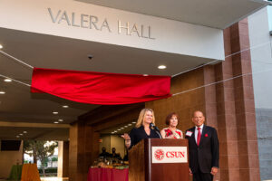 outdoors, the building signage reading "Valera Hall" is unveiled. President Beck, Debbie Valera and Milt Valera stand at a podium beneath the sign.