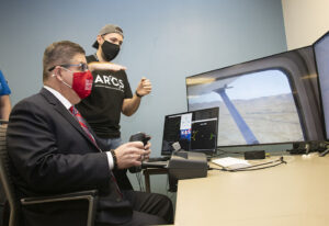 Chancellor Joseph Castro sits at a computer console, holding the controls of an autonomous flight simulator program. CSUN student Sarkis Mikaelian stands behind him, giving directions.