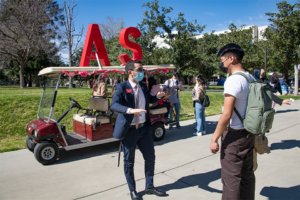 Student in suit speaks to other student. Decorated Associated Students golf cart in background.