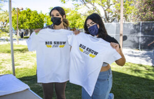 Two young women wearing masks display white t-shirts with "Big Show 2022" written on them