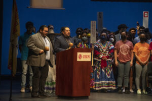 Two men stand at podium on stage with students in background