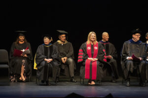 CSUN President Erika D. Beck in black academic robe with red sash with other academics in black robes on black floored stage