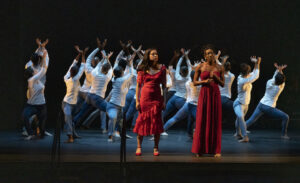 Two women wearing red dresses in front of group of dancers wearing white shirts and blue pants on dark stage.