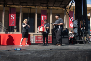 Erika D. Beck on stage being handed a box by one man while another man looks on