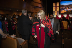 CSUN President Erika D. Beck leads the recessional, walking out of The Soraya's Great Hall after the investiture ceremony. She is wearing a red and black blanket presented by the Fernandeño Tataviam Band of Mission Indians. Audience members stand and clap.