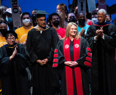 President Erika D. Beck stands on stage at The Soraya, in a black and red CSUN academic gown, and flanked by President Emerita Jolene Koester, David Nazarian, Robert Taylor and CSU Acting Chancellor Steve Relyea. Behind her is a crowd of CSUN students in colorful T-shirts.