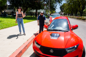 Narrow red car in foreground with two people standing on sidewalk looking at the car.