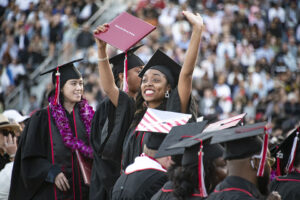 A new graduate in cap and gown holds her red diploma cover in the air and waves toward the bleachers, at Commencement 2022.
