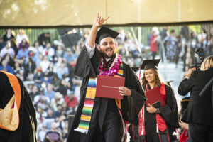 A graduate in cap, gown and sash crosses the Commencement stage, holding his red diploma cover and signing "I Love You" in ASL.