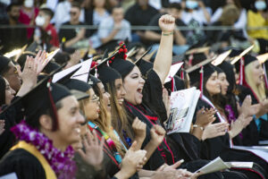A row of graduates in caps, gowns, sashes and flower leis clap and cheer, at Commencement 2022. One graduate raises her fist in the air, in triumph.
