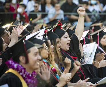 A row of graduates in caps, gowns, sashes and flower leis clap and cheer, at Commencement 2022. One graduate raises her fist in the air, in triumph.
