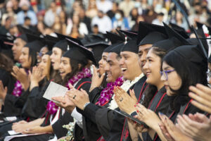 A row of graduates in caps, gowns, sashes and flower leis clap and cheer, at Commencement 2022.
