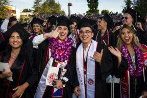 A row of graduates in caps, gowns, sashes and flower leis stand and smile, celebrating at Commencement 2022.
