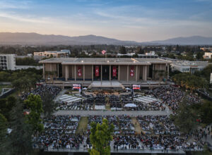A drone camera aerial view of the University Library and Commencement crowds in bleachers and sections of chairs, with the CSUN campus and north San Fernando Valley beyond.