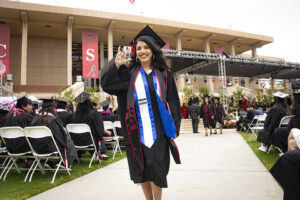 A CSUN graduate from the class of 2021, in cap and gown, signs "I Love You" in ASL, as she walks in front of the Commencement stage.