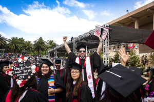 A crowd of graduates in caps, gowns and sashes smile and raise their arms in celebration, in front of the University Library at Commencement 2022.