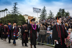 Graduates in caps and gowns march in to a Commencement ceremony, as one carries a sign that reads: Electrical & Computer Engineering. They are walking in front of a metal barricade, with a large crowd in the bleachers behind them.