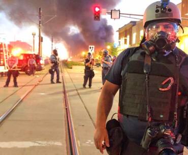 Man stands on street with fire behind him. He is wearing riot helmet and gas mask.