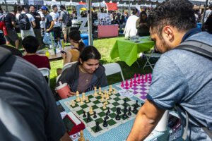 Students sit at table with two chessboards in front of them. One person stands on other side of boards, leaning over them.