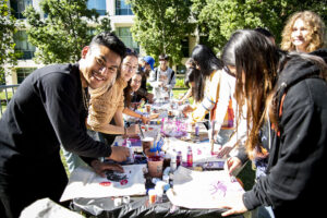 Students stand on both sides of a table that has paint and paper laid out.