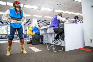 Student wearing red kerchief on his head, boots and blue shirt stands in the library with students studying in the background.