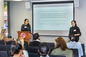 Two women stand, one at podium, in front of a screen. Seated audience members are in the foreground.