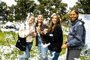 A group of four students covered in snow hold their cups of hot cocoa and smile for the photo.