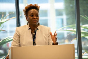 Woman in white blazer standing behind podium with her hand up mid-speech.