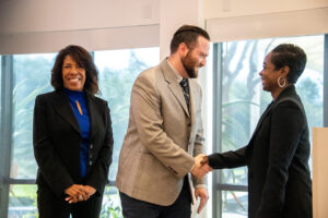 Man shaking hands with woman on the right, while woman on the left stands with hands crossed and is smiling.