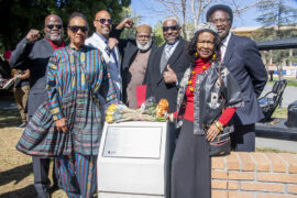 Group of 7 people stand behind metal plaque mounted on concrete base.
