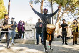 Man stands wearing drum with arms lifted and people behind him.