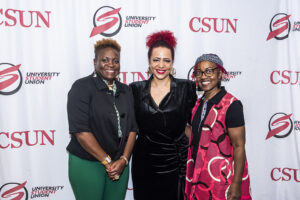 Three women pose, smiling, in front of CSUN banner
