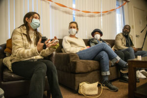 Student sitting on a chair speaks while guests nearby sitting on a couch look and listen.