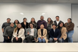 Fourteen of the teams researchers are posing for a picture . The group is organized into two lines while positioned in front of a whiteboard, and they are all wearing neutral and muted colors.