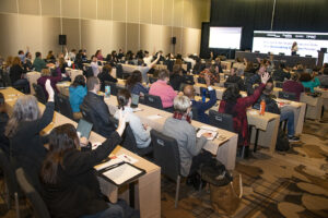 People seated in conference room facing a stage filled with video monitors