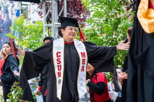 Diana Vicente, in cap and gown and white sash, holds her arms out wide as she crosses the commencement stage.