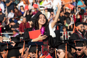 Graduate in cap and gown, clutching her diploma cover waves from the crowd